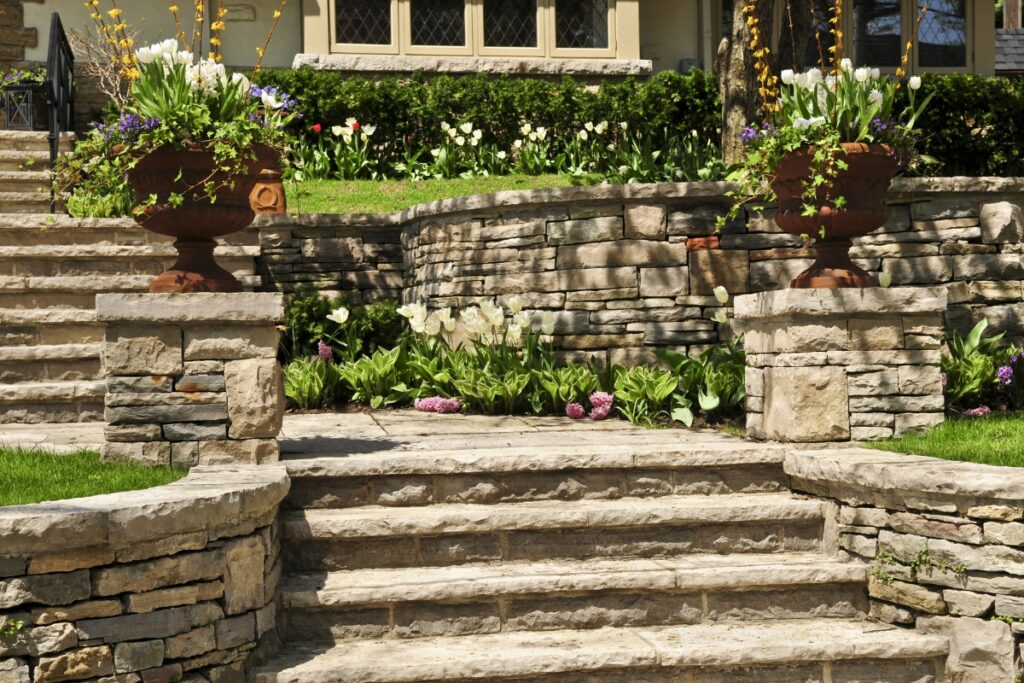 A garden stone border surrounds the steps leading up to a house adorned with flowers in pots.