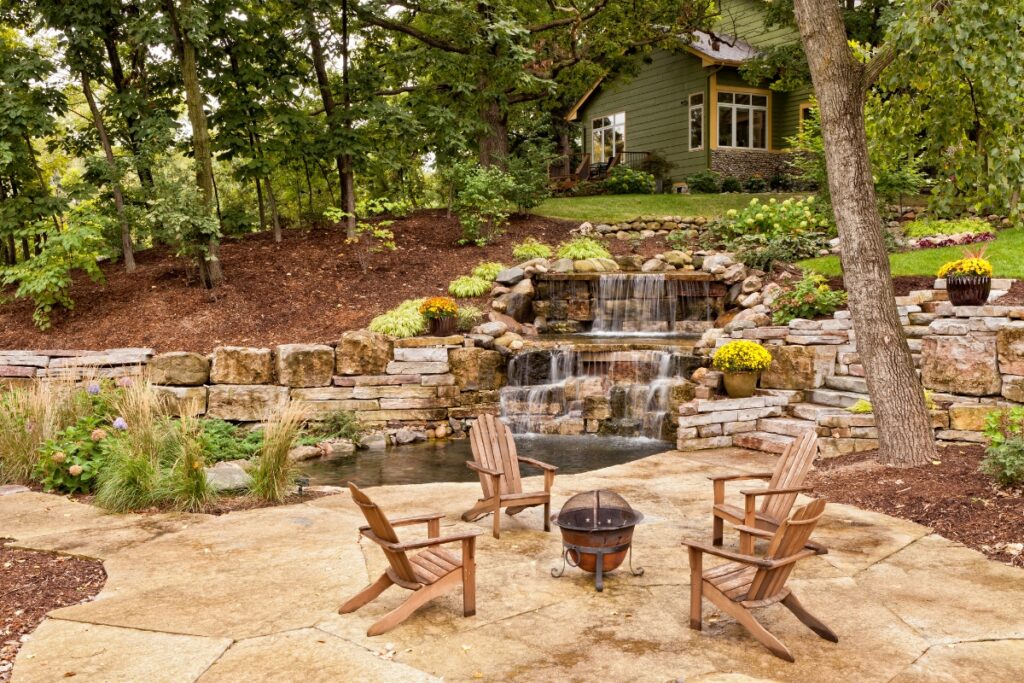 Backyard patio with a fire pit and wooden chairs overlooking a landscaped waterfall and pond, embodying current outdoor living trends.