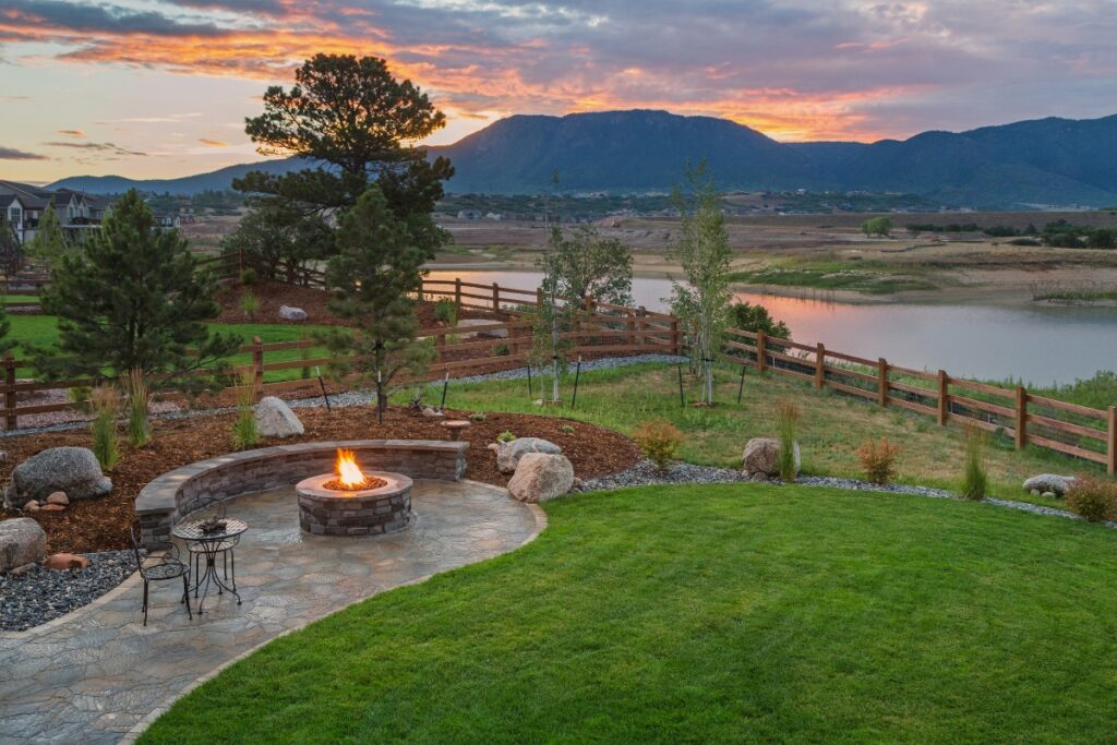 A landscaped backyard, reflecting the latest outdoor living trends, with a fire pit and seating area overlooking a lake at sunset.