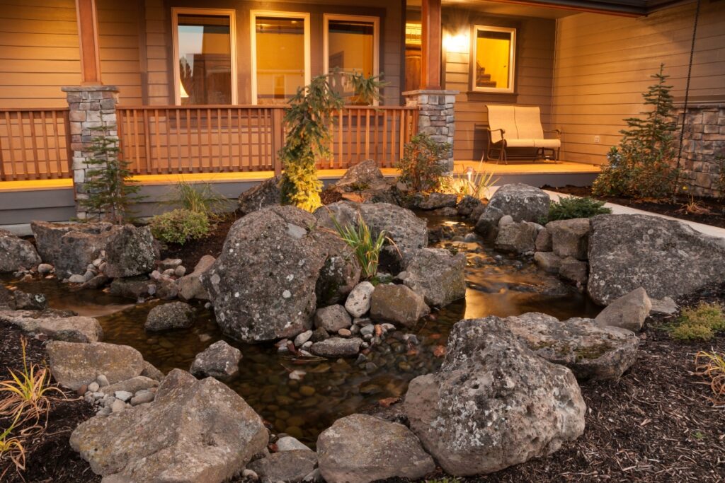 A tranquil pond with large stones in front of a house with a cozy porch at dusk, embodying the latest outdoor living trends.