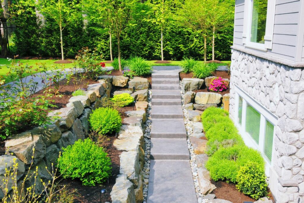 Stone staircase leading through a vibrant garden featuring eco-friendly landscaping next to a house with stone siding, surrounded by lush greenery and flowers.
