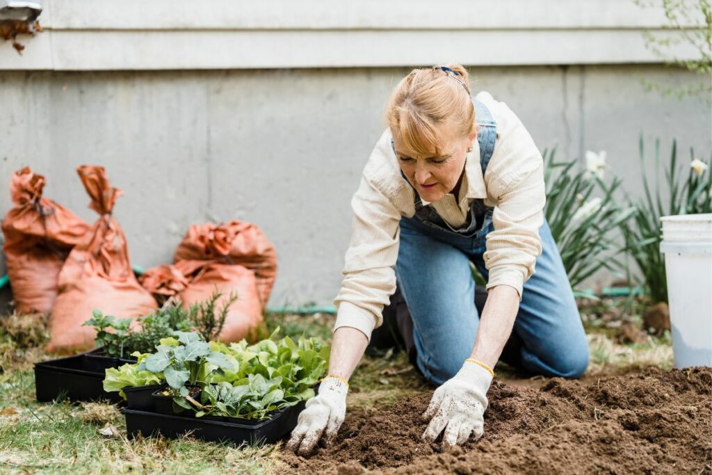 A woman plants seedlings in a garden, practicing eco-friendly landscaping by kneeling in the soil with gardening gloves, a trowel, and plants nearby.