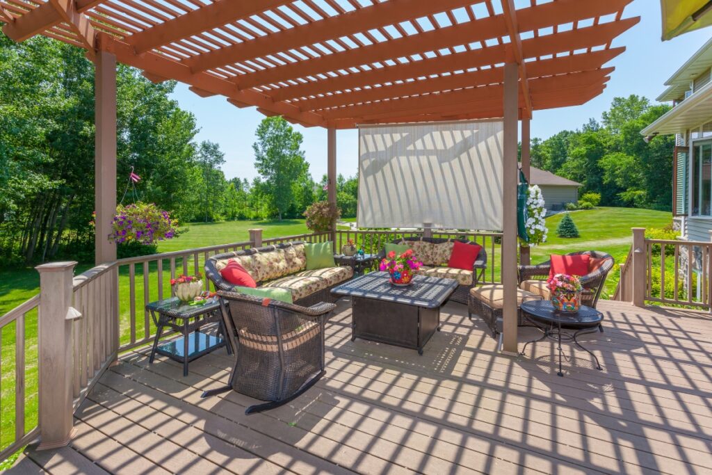 Sunny backyard deck with hardscape design including a pergola, comfortable seating, and blooming flowers on a clear day.