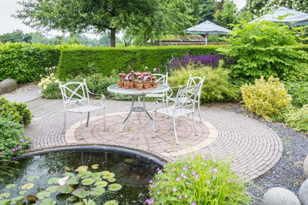 A garden patio area featuring a hardscape design with a table and chairs set near a pond with water lilies.
