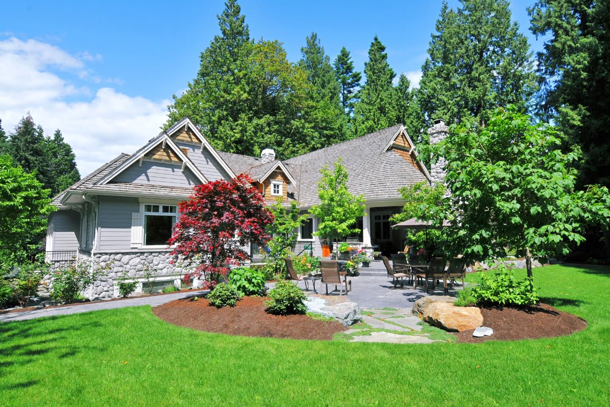 A grey stone and wood house with a gabled roof is surrounded by a lush green lawn, various trees, shrubs, and a paved patio area with outdoor seating. The meticulous landscaping significantly enhances the property value.