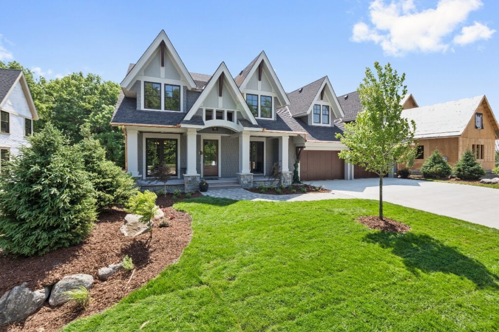 A modern two-story house with a well-maintained lawn, landscaping that boosts property value, including shrubs and trees, and a two-car garage. The house features a peaked roof with multiple gables and a stone walkway.