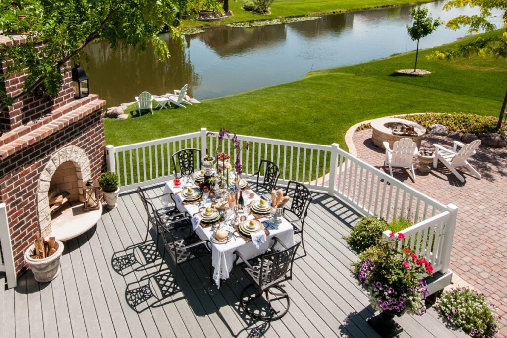 A patio deck with a dining table set for a meal overlooks a lawn and a serene river. Nearby are Adirondack chairs by a fire pit, various potted plants, and well-designed landscaping that enhances the property's value.