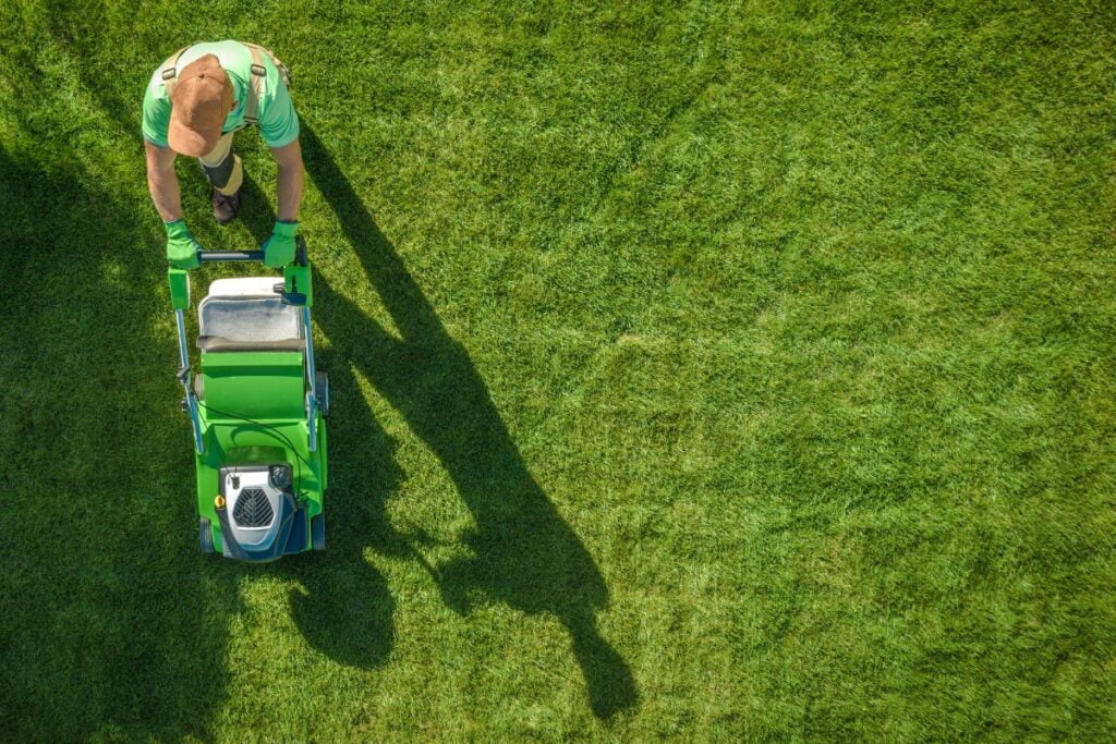 A person, seen from above, wearing green work gloves and pushing a green lawnmower on a neatly cut grassy lawn, skillfully enhancing the landscaping to boost the property's value.