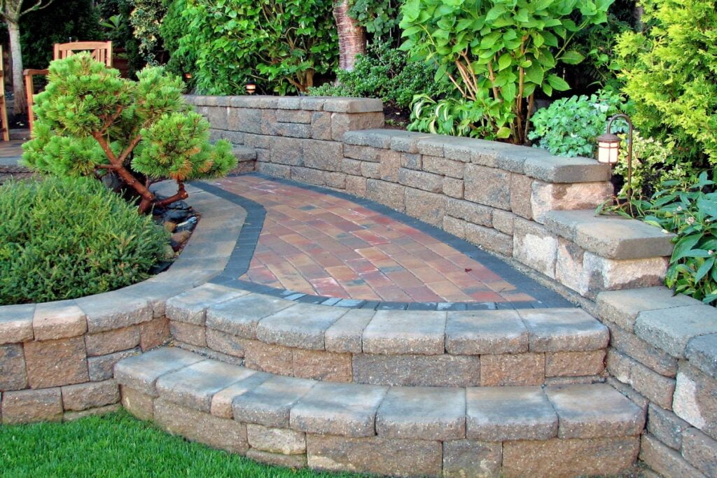 A garden path with red and black bricks is flanked by raised stone flower beds and greenery, enhancing the landscaping and boosting the property value while creating a serene, landscaped area.