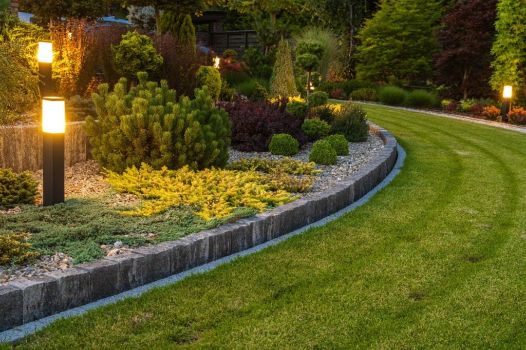 A well-maintained garden with a curved stone border, various plants, and illuminated pathway lights next to a grassy lawn enhances both the landscaping and property value.