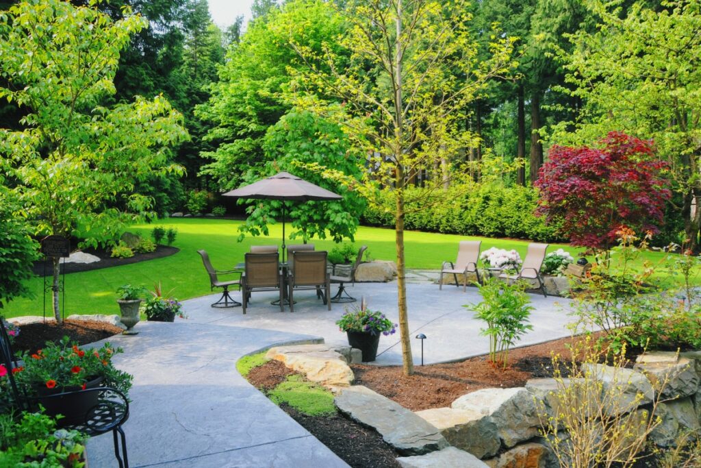 Lush wildlife-friendly backyard garden featuring a patio with table and chairs under a sun umbrella, surrounded by vibrant greenery and flowers.