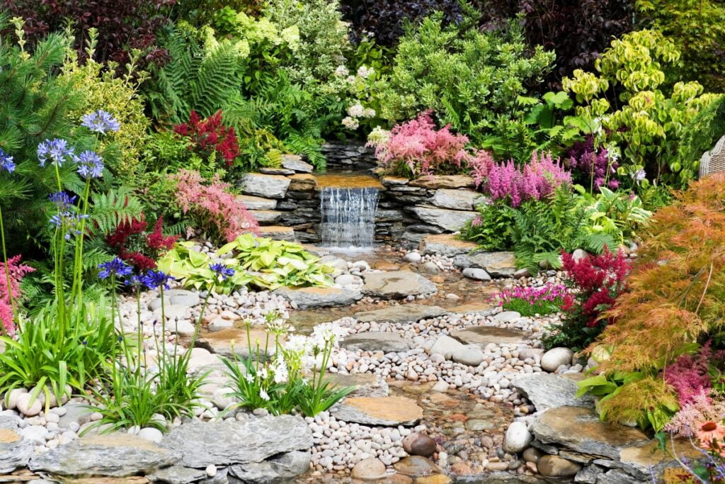 A vibrant, wildlife-friendly garden with a small waterfall and a stone-lined stream, surrounded by various colorful flowers and plants.