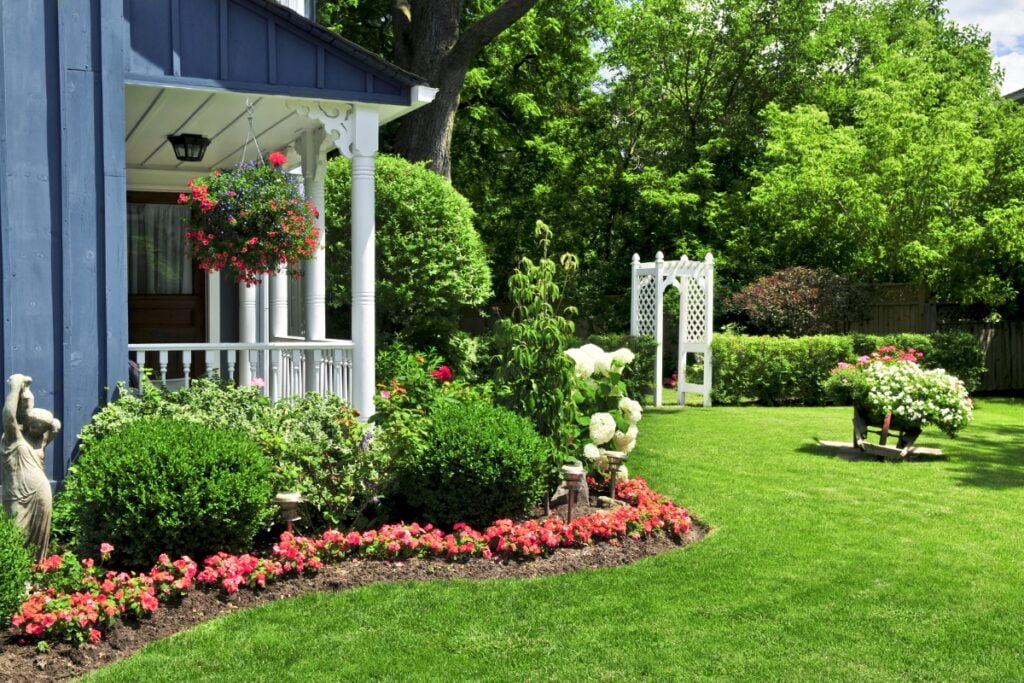 Lush garden designed for wildlife-friendly landscaping, featuring a vibrant display of flowers near a blue house with a white porch, including a statue and a trellis under a clear sky.