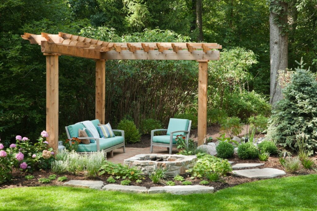 A wooden pergola with teal furniture and potted plants underneath is the centerpiece of this Atlanta outdoor living space, surrounded by a garden with various green plants and stone walkways.