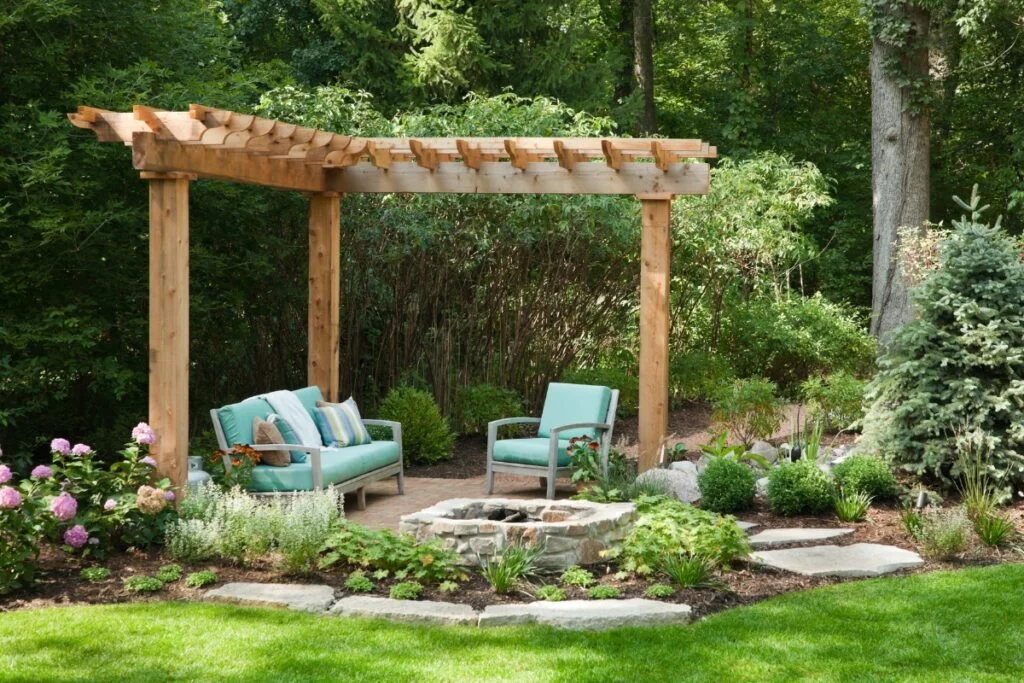 Atlanta Outdoor Living Spaces Transformed into Tranquil Backyard Retreats 1 A wooden pergola with teal furniture and potted plants underneath is the centerpiece of this Atlanta outdoor living space, surrounded by a garden with various green plants and stone walkways.