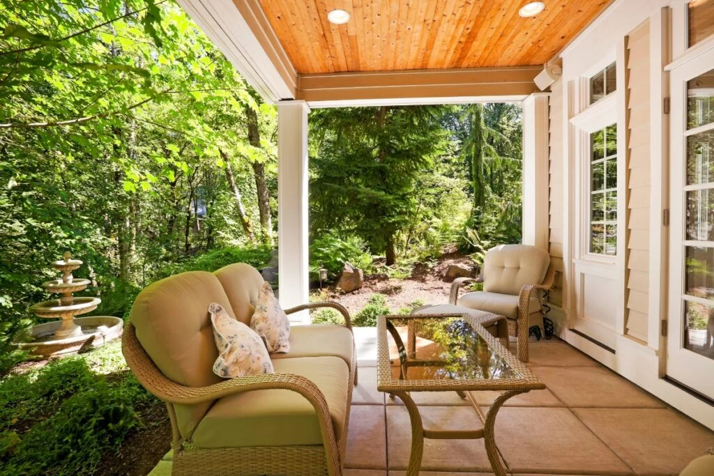Atlanta Outdoor Living Spaces Transformed into Tranquil Backyard Retreats 2 A covered patio with beige wicker furniture and cushions overlooks a lush garden with a stone fountain. The ceiling is wood-paneled and recessed lighting is installed, exemplifying the charm of Atlanta outdoor living spaces.