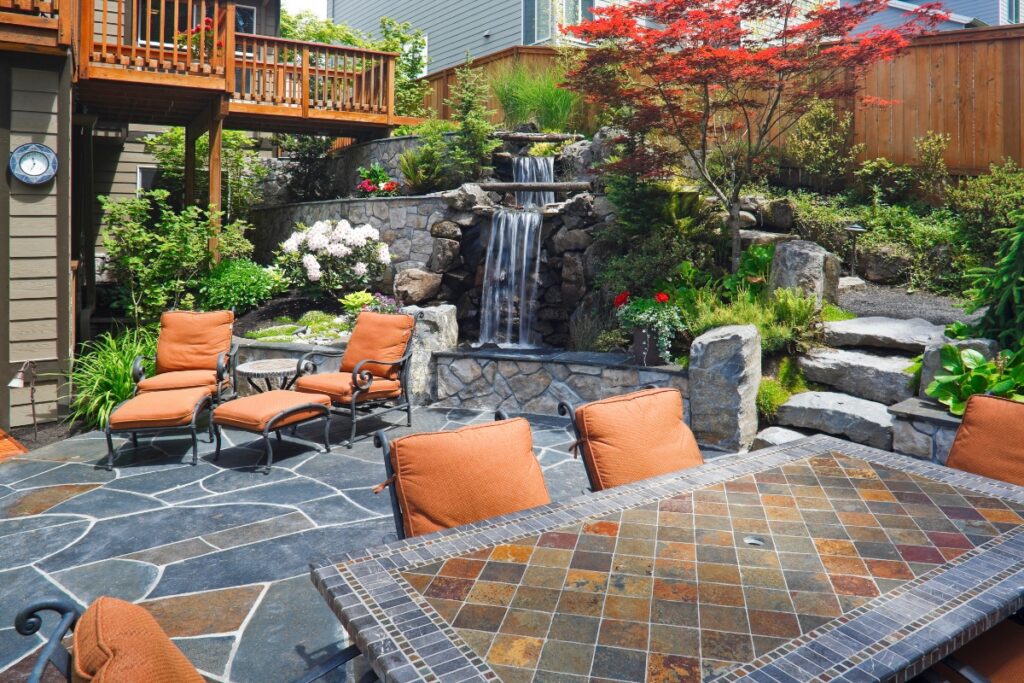 A backyard patio with cushioned chairs and a stone table overlooks a small waterfall and lush garden with a variety of plants, epitomizing Atlanta outdoor living spaces. Stone steps lead up to another level.