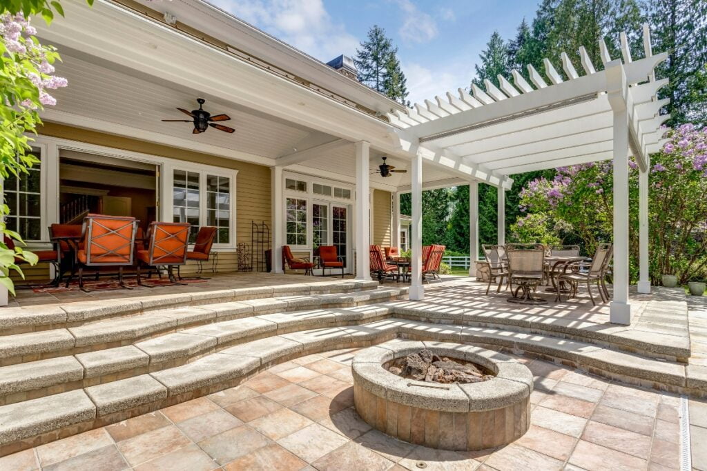 Spacious outdoor patio with a pergola, multiple seating areas, a fire pit, and steps leading to the house with large glass doors, surrounded by lush greenery—a perfect example of Atlanta outdoor living spaces.