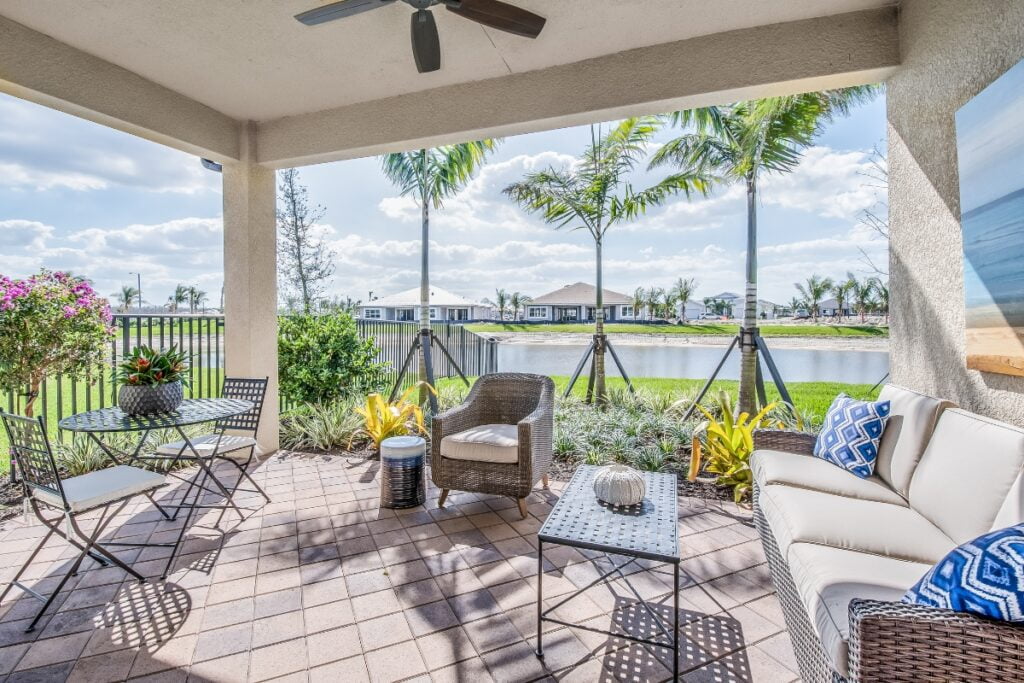 A covered patio with seating includes a wicker sofa, armchair, and a table set for two, overlooking a pond and modern houses in the background. Palm trees and potted plants enhance the serene view of one of Atlanta's finest outdoor living spaces.