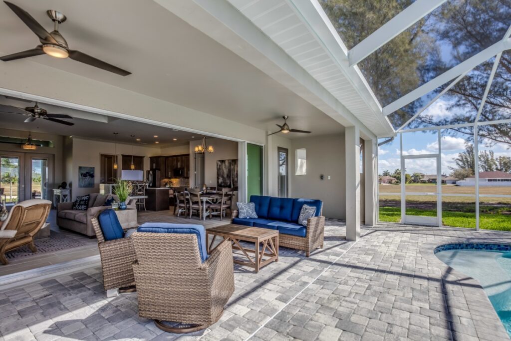 A covered outdoor patio in one of Atlanta's finest outdoor living spaces features wicker furniture with blue cushions, a dining area, ceiling fans, and a view of a pool under a screened enclosure, adjoining an open-concept living and kitchen area.