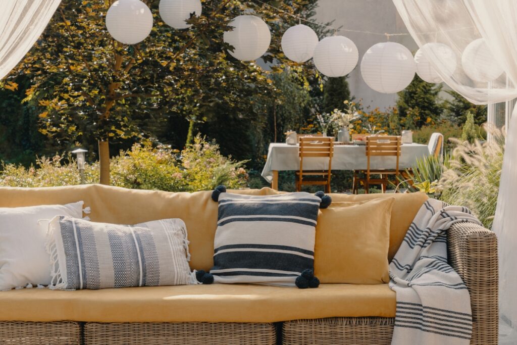 A wicker couch with striped and solid pillows is set up outdoors under white lanterns, epitomizing Atlanta outdoor living spaces. In the background, a dining table adorned with a light tablecloth and floral arrangements creates an inviting ambiance, surrounded by lush greenery.