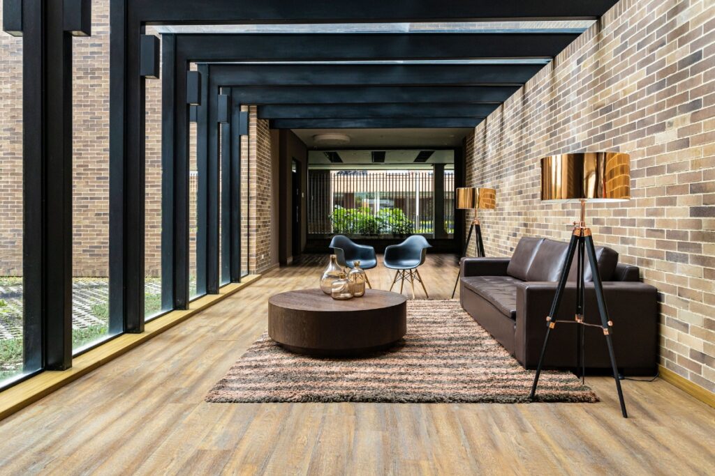 A modern lounge area with a brown sofa, round coffee table, two black chairs, two floor lamps, and brick walls features large windows on the left side that let in ample natural light, reminiscent of Atlanta outdoor living spaces.