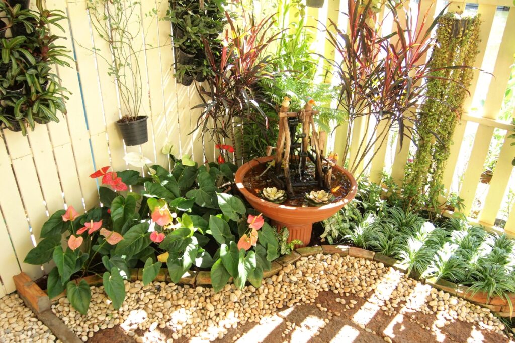 A small garden corner with various potted plants, a fountain in the center, decorative rocks on the ground, and a white picket fence backdrop offers an idyllic setting to try out some summer gardening tips.