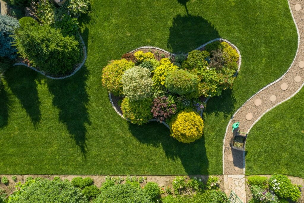 Aerial view of a well-maintained garden featuring a curved path with stepping stones, lush green lawn, and a central colorful flower bed. A person with a wheelbarrow is walking on the path, embodying summer gardening tips in action.