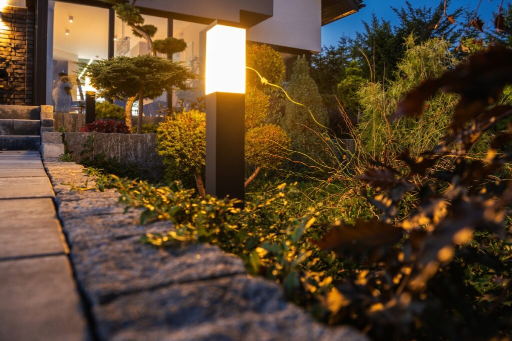 Glowing outdoor light illuminating a garden path with plants and shrubs near a modern house during dusk, perfect for soaking in the ambiance and discovering summer gardening tips.