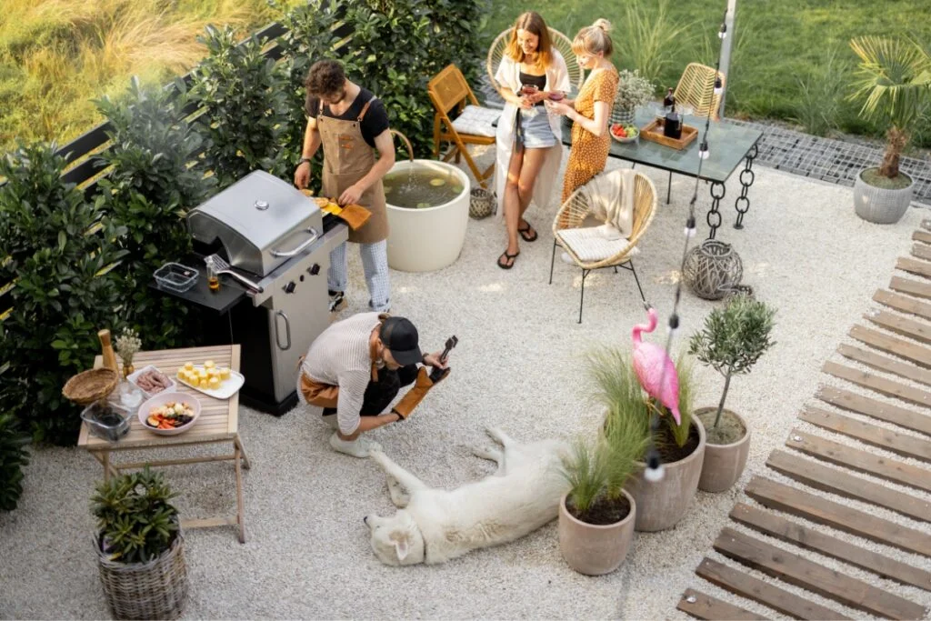 Outdoor Hosting Ideas to Create Stunning Backyard Entertainment Areas 9 Four people are enjoying a barbecue in a garden with a grill and outdoor furniture, showcasing great outdoor hosting ideas. One person is petting a large white dog lying on the ground. Plants and a pink flamingo decoration surround the scene.