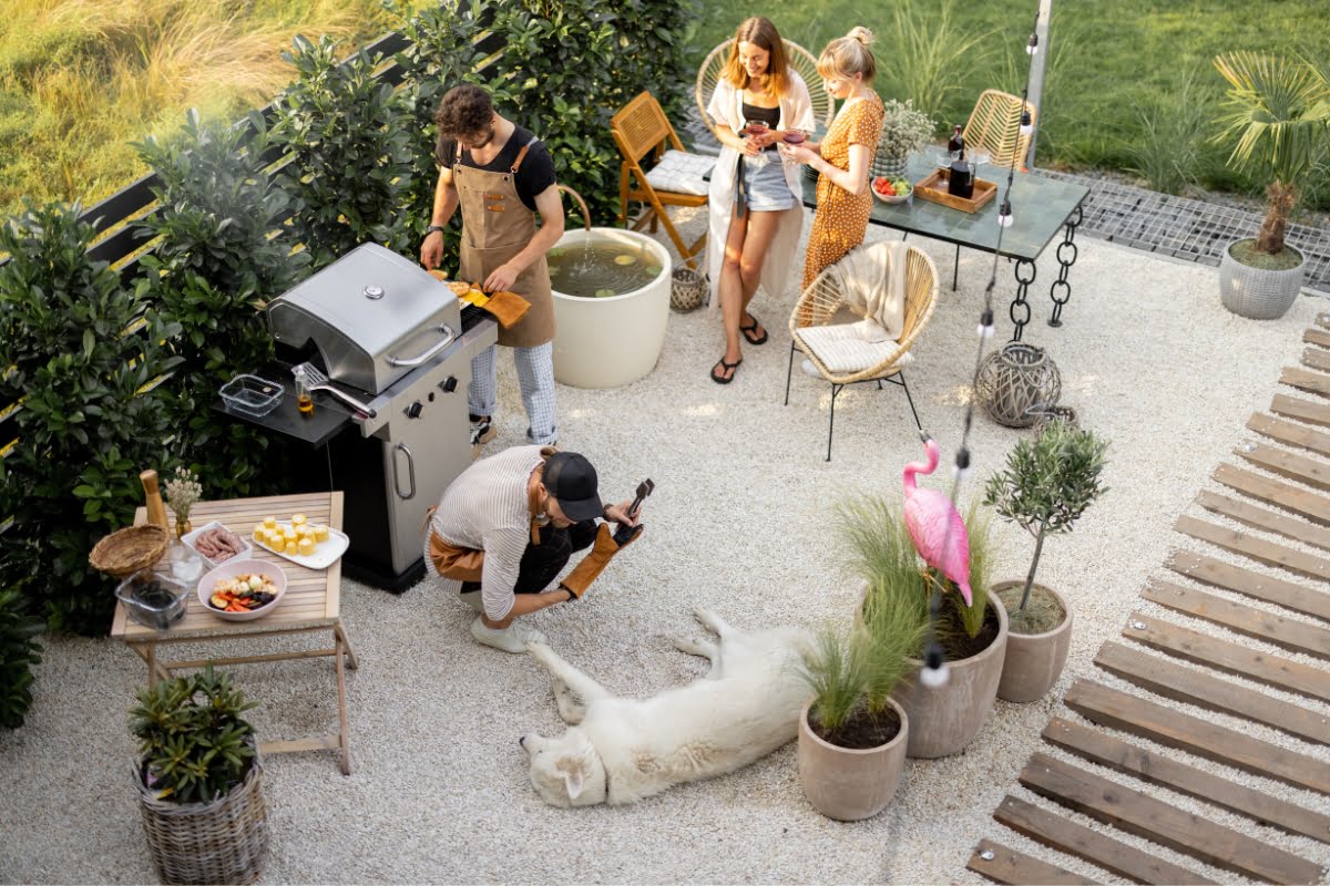 Four people are enjoying a barbecue in a garden with a grill and outdoor furniture, showcasing great outdoor hosting ideas. One person is petting a large white dog lying on the ground. Plants and a pink flamingo decoration surround the scene.