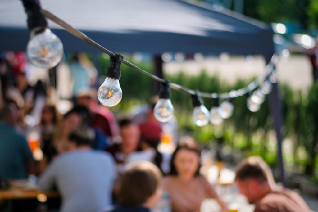 Outdoor hosting ideas come to life with people sitting at tables under a shaded canopy, a string of clear light bulbs twinkling in the foreground.