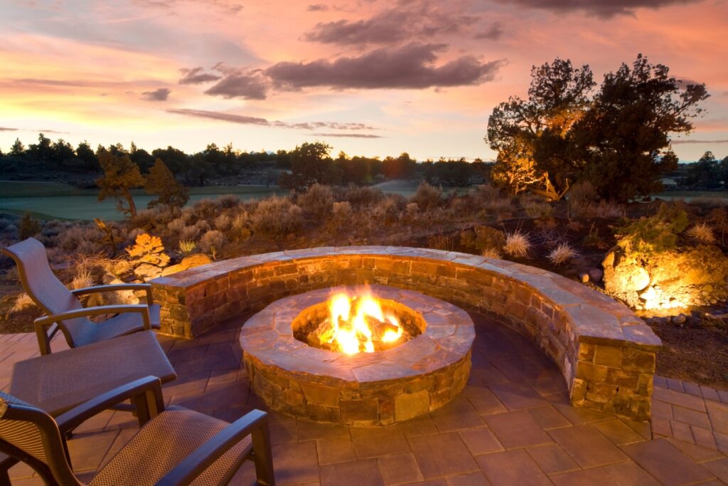 Outdoor seating area with a circular stone fire pit at sunset, perfect for outdoor hosting ideas. The scene is surrounded by trees and shrubs, with a clear sky in the background.