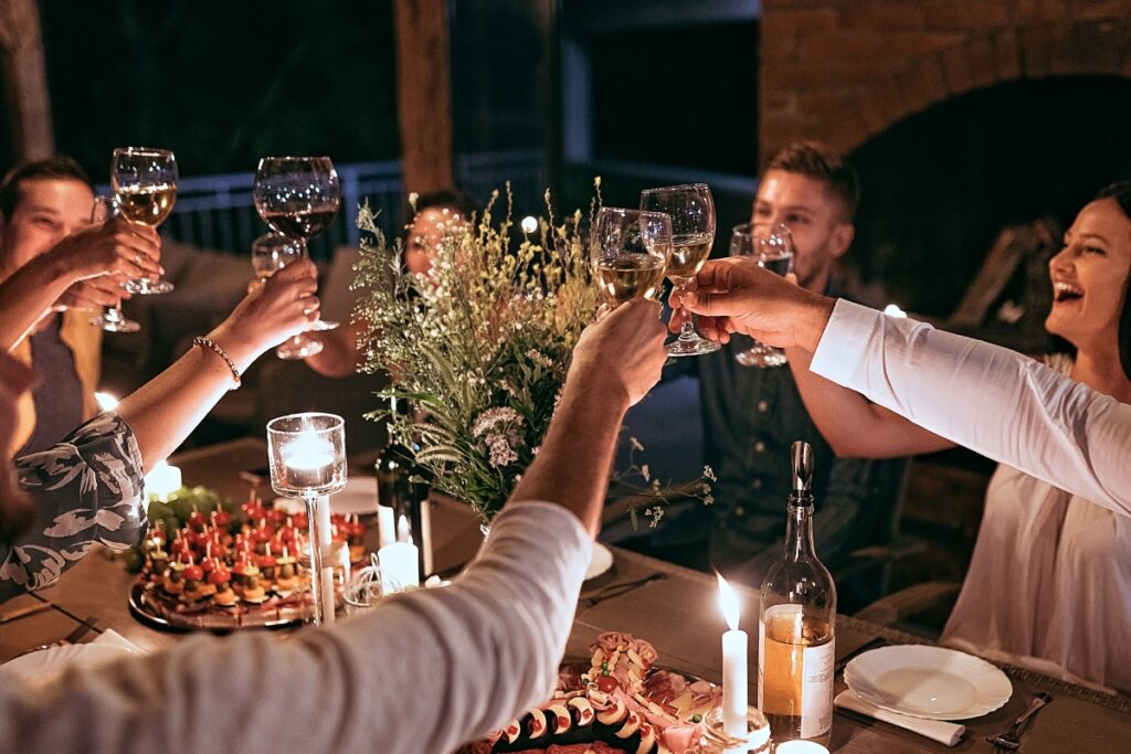 A group of people clinking wine glasses in a toast around a candlelit dinner table with flowers, food, and drinks—a perfect setting for outdoor hosting ideas.