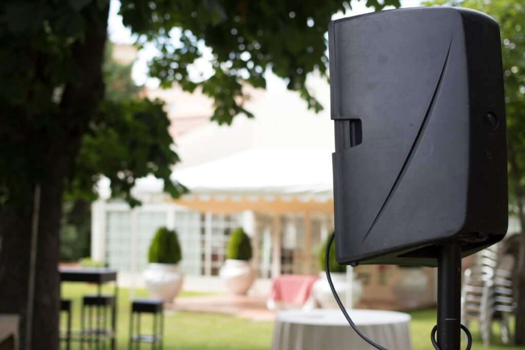 A black speaker on a stand is set up outdoors in a garden with trees and a white tent in the background, showcasing one of many great outdoor hosting ideas.