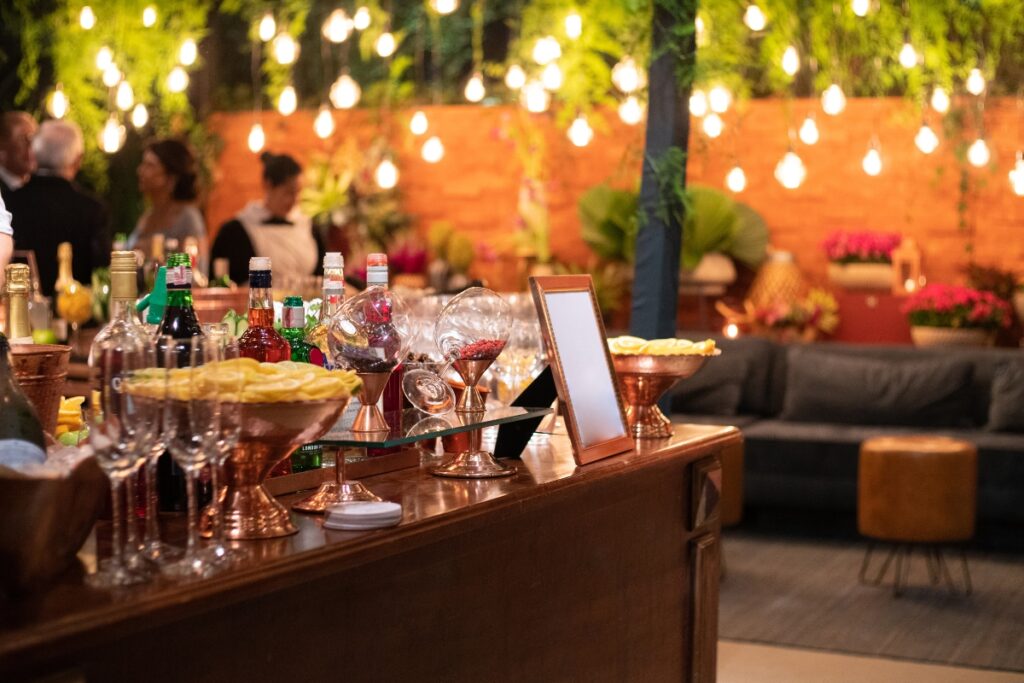 A bar counter with various drinks and glasses in an indoor space, with several people enjoying themselves and hanging fairy lights in the background—perfect for inspiring outdoor hosting ideas.