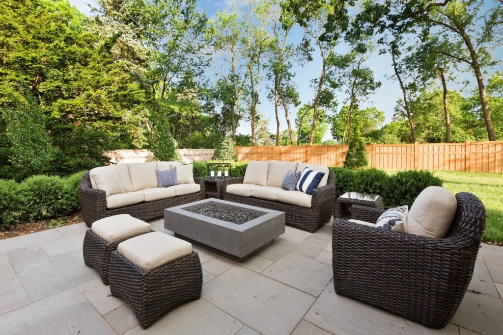 An outdoor patio with wicker furniture, including two sofas, two stools, a side chair, and a concrete fire pit table—perfect for outdoor hosting ideas. The patio is surrounded by lush greenery and a wooden fence.