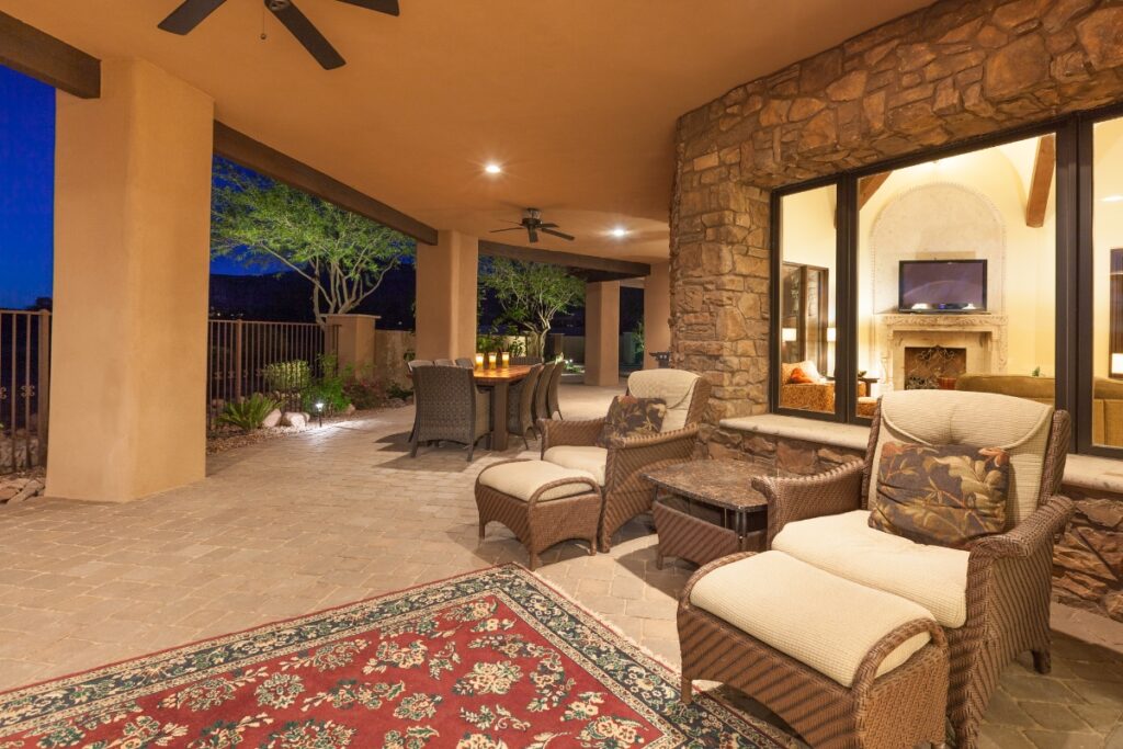 Covered outdoor patio at dusk with wicker lounge chairs, a dining table, ceiling fans, and a patterned rug. Large windows reveal an interior room with a lit fireplace and television—perfect for gathering some outdoor hosting ideas.