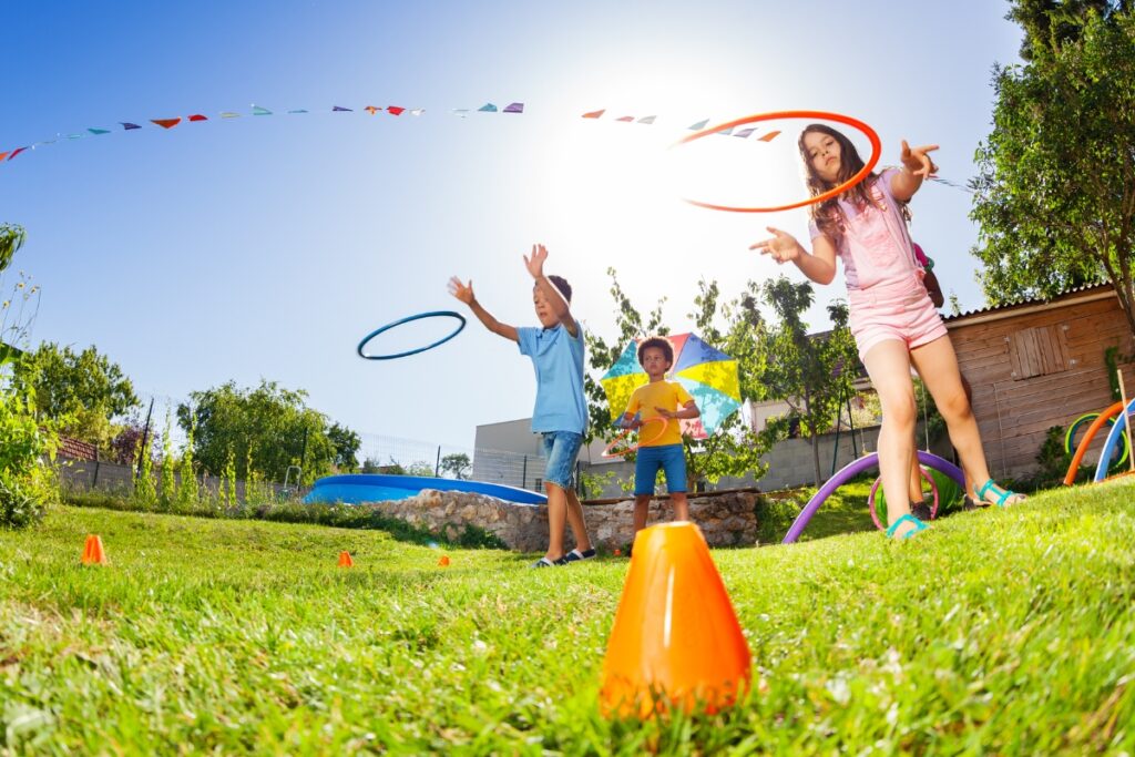 Three children playing with hula hoops outdoors on a grassy area under a sunny sky, with decorative flags hanging in the background—perfect inspiration for outdoor hosting ideas.