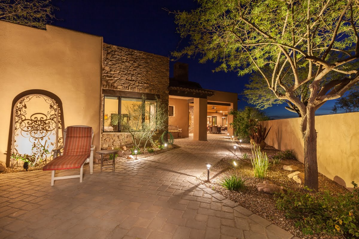 A modern house with stucco walls and stone accents features a lit walkway with elegant outdoor lighting, a decorative iron gate, outdoor seating, and desert landscaping under a clear night sky.