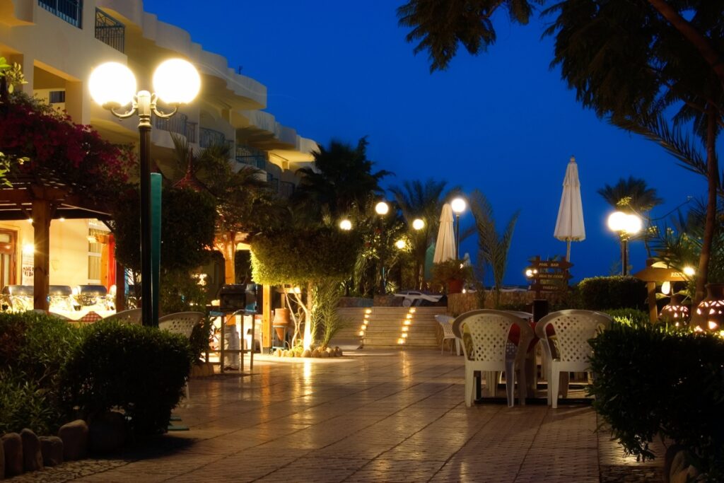 Outdoor restaurant with white chairs and umbrellas under soft outdoor lighting at night. Lit street lamps add to the ambiance, while a building with balconies and palm trees provides a charming backdrop.