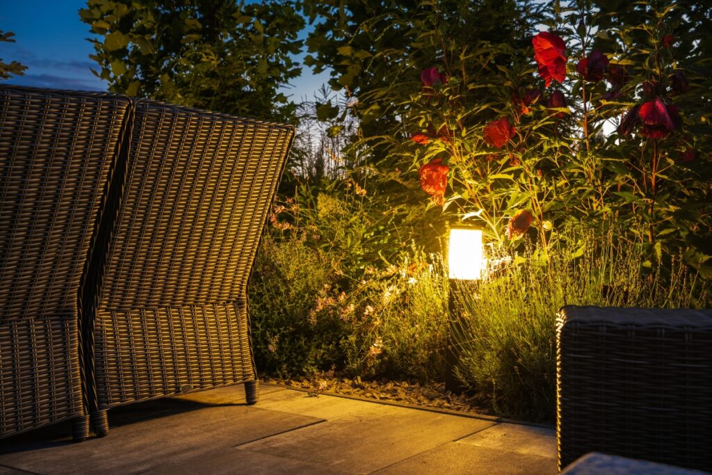 Outdoor seating area with wicker furniture and garden plants under evening outdoor lighting. A small lamp illuminates red flowers and greenery nearby.