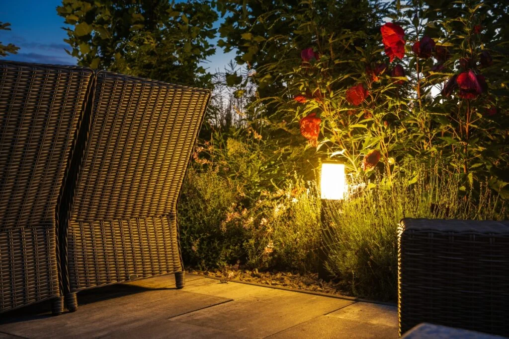 How Outdoor Lighting Elevates Your Home's Aesthetic and Security 3 Outdoor seating area with wicker furniture and garden plants under evening outdoor lighting. A small lamp illuminates red flowers and greenery nearby.