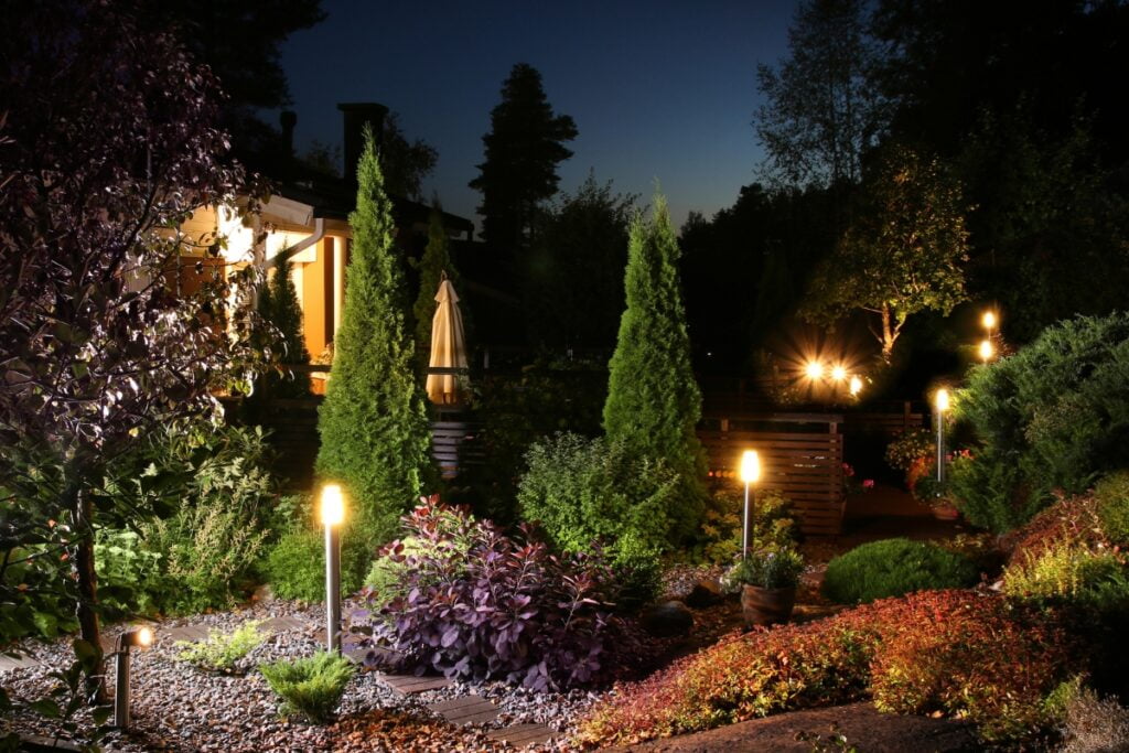 A well-lit garden at dusk featuring various plants, shrubs, and trees bordered by illuminated pathways with outdoor lighting and a house with glowing windows in the background.