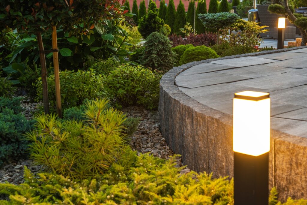 A landscaped garden adorned with outdoor lighting features illuminated pathway lights surrounding a circular stone patio. Numerous types of plants and shrubs are thoughtfully arranged around the area.