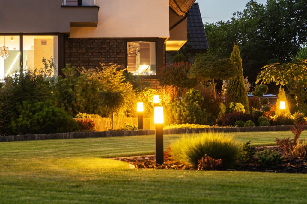 A residential house with outdoor lighting illuminating the garden, a well-maintained lawn, and landscaped greenery at dusk.