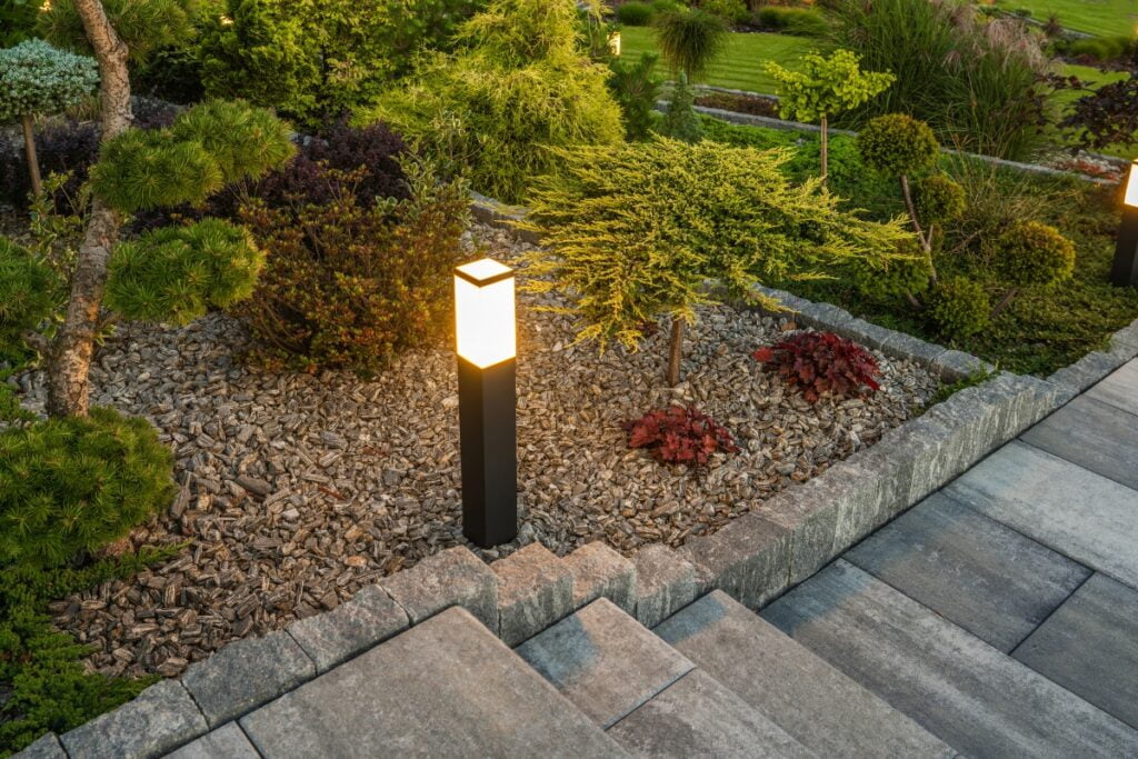 Illuminated garden path light next to a stone walkway and stairs, surrounded by various plants and mulch, brings charm and functionality to your outdoor lighting setup.