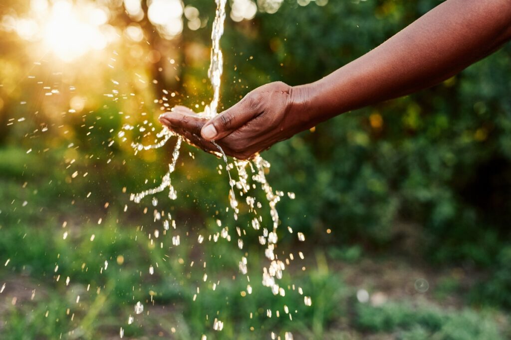 A person is holding cupped hands under a stream of water outside, with sunlight and greenery in the background, embracing sustainable home practices.