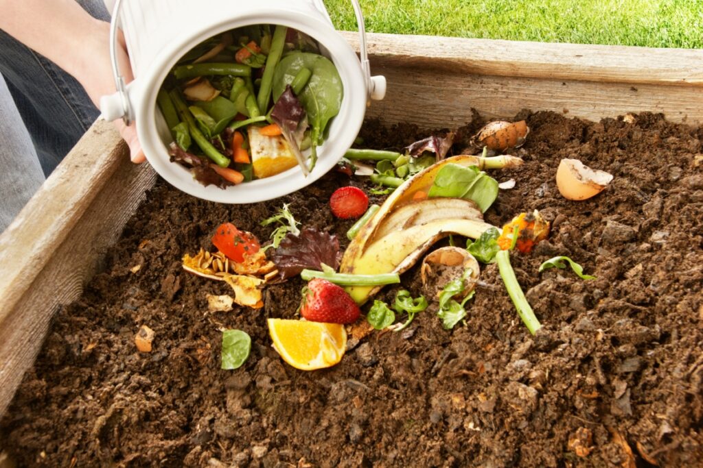 A person embraces sustainable home practices by dumping a bucket of food scraps, including vegetables and fruit peels, onto a compost pile in a wooden container.