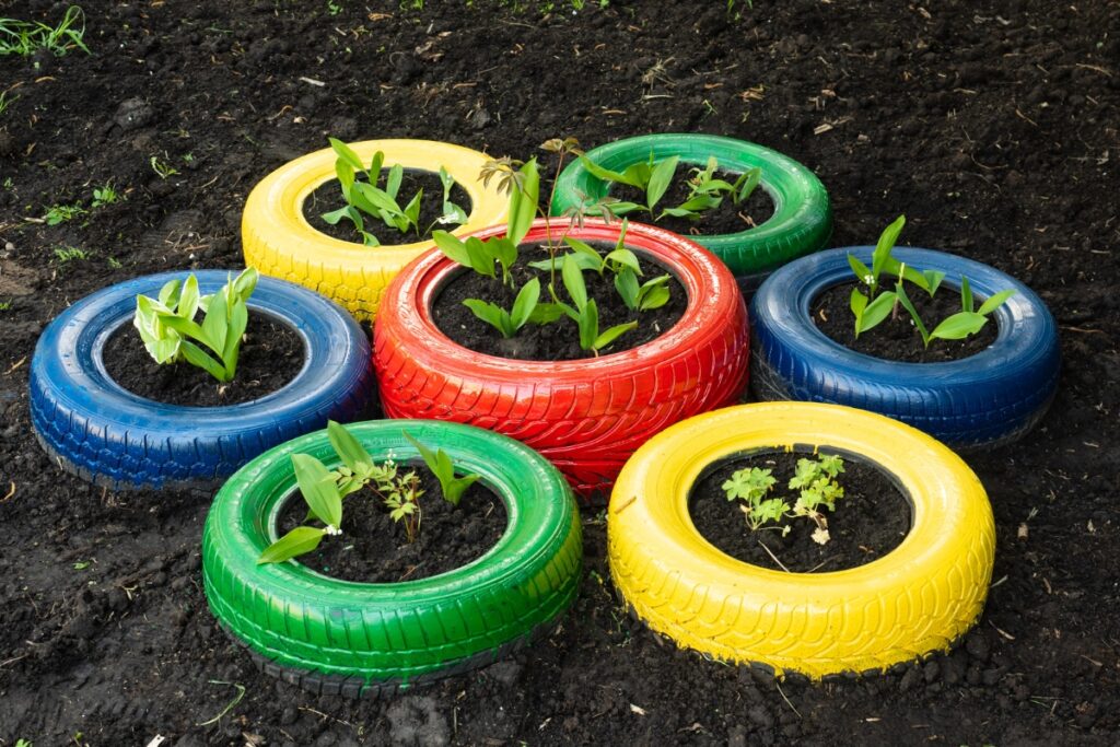 Colorful tires painted red, blue, yellow, and green are arranged in a circle and transformed into planters filled with young plants growing in soil, showcasing sustainable home practices.